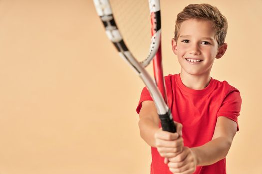 Adorable male child tennis player holding racket and smiling while standing against light orange background