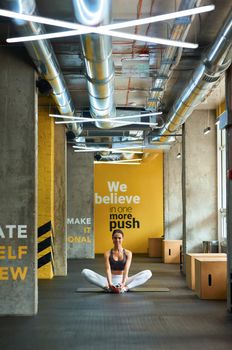 Stretching exercises. Vertical shot of a young fitness woman in sportswear sitting on yoga mat at gym and warming up before workout. Sport, wellness and healthy lifestyle