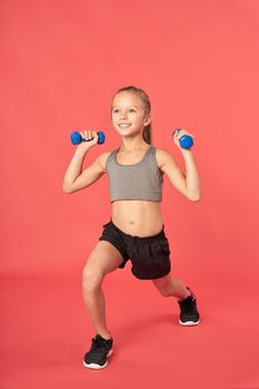 Adorable female child in sportswear holding dumbbells and smiling while doing exercise against red background