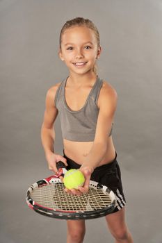 Adorable girl tennis player with racket and ball looking at camera and smiling while standing against gray background