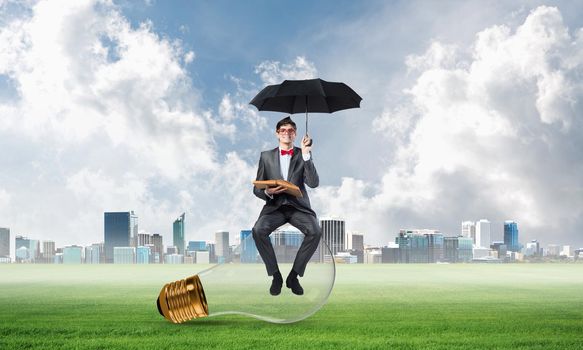young businessman with an umbrella and a book, sitting on a light bulb. concept of finding new ideas in business