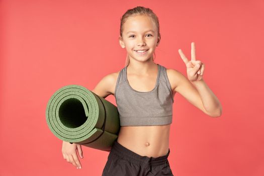 Cute female child with fitness exercise mat looking at camera and smiling while standing against red background