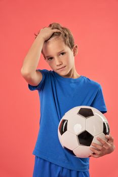 Adorable male child in blue shirt holding football ball and fixing hair while looking at camera with serious expression