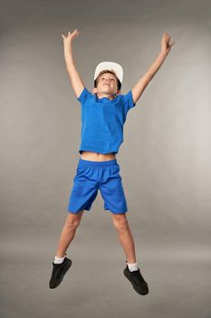 Cute sporty boy wearing blue shorts and cap while raising hands and jumping in the air