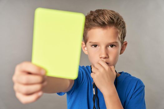 Adorable male child showing penalty card and using referee whistle while standing against gray background