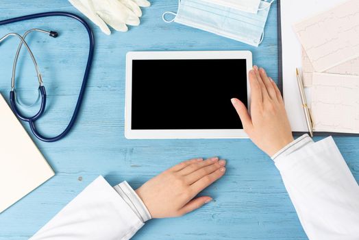 Top view of doctor hands with tablet computer. Cardiac examination in hospital. Therapist sitting at blue wooden desk with stethoscope, medical gloves and mask. Examination and consultation in clinic