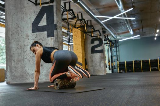 Rear view of a young beautiful sportive woman in sportswear exercising with foam roller on yoga mat at gym, using special fitness equipment. Sportive people, training and workout concept