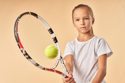 Cute female child tennis player hitting the ball with racket while standing against light orange background