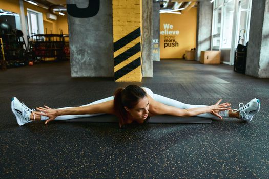 Young flexible fitness woman in sportswear sitting on the floor at gym and practicing transverse twine, stretching muscles after workout. Sport and healthy lifestyle concept