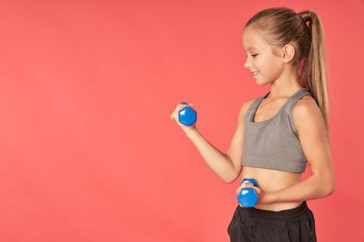 Adorable female kid in sportswear holding dumbbells and smiling while standing against red background
