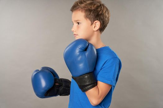 Serious male child boxer wearing sports boxing gloves and blue shirt while doing fighting stance