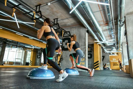 Full length of two young athletic women in sportswear exercising together with resistance fitness bands at crossfit gym, doing squats and training legs. Workout, sport, wellness and healthy lifestyle