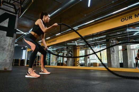 Cardio workout. Young strong athletic woman wearing sport clothes exercising with battle ropes at crossfit gym, full length. Sport, training and healthy lifestyle