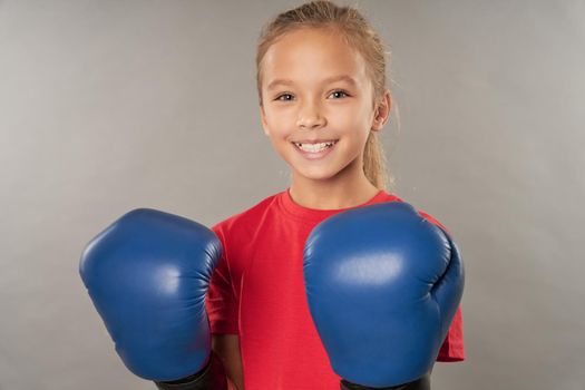 Adorable female child boxer wearing boxing gloves and red shirt while looking at camera and smiling