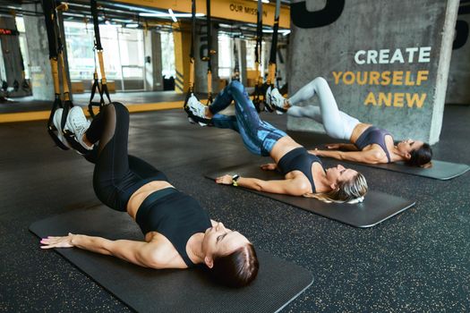 Full body training. Three young caucasian athletic women in sportswear lying on the floor and exercising with fitness trx straps at gym. Sport, people working out, wellness and healthy lifestyle