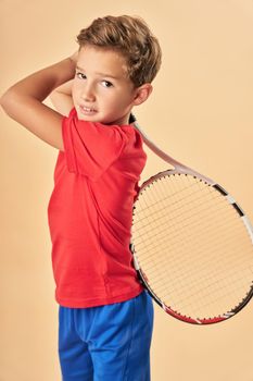 Adorable male child in sportswear looking at camera and smiling while holding tennis racket