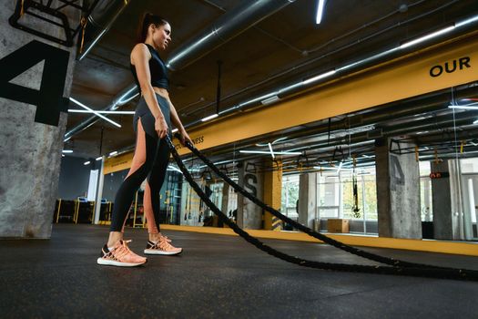 Strength workout. Young strong athletic woman exercising with battle ropes at crossfit gym, full length. Sport, training and healthy lifestyle