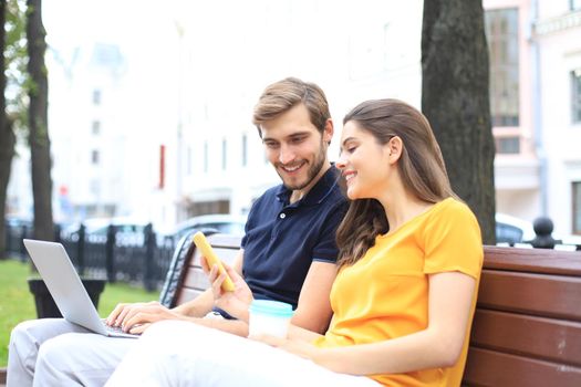Attrative young couple using laptop computer while sitting on a bench outdoors, holding mobile phone