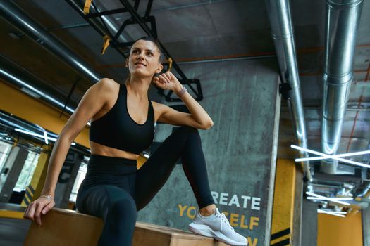 Resting after workout. Young beautiful athletic girl in sportswear sitting on wooden crossfit jump box at gym and looking aside. Sportive people, training, healthy lifestyle concept