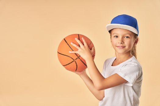 Charming female child basketball player holding game ball and smiling while standing against light orange background