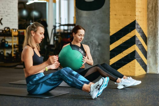 Staying active. Two young caucasian athletic women in sportswear exercising with fitness ball at gym, working out together. Sport, training, wellness and healthy lifestyle