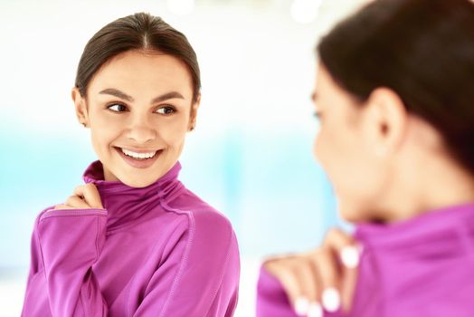 Close up portrait of a young beautiful caucasian woman, female fitness instructor in sportswear looking in the mirror and smiling while standing in studio or gym. Sport, wellness and healthy lifestyle