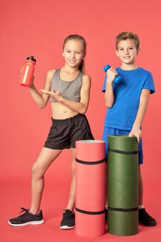 Adorable female child with bottle of water looking at camera and smiling while boy holding dumbbell and yoga mat