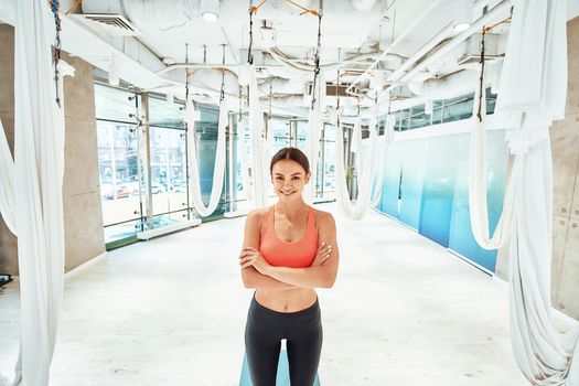 Young attractive and happy woman in sportswear, female fitness instructor keeping arms crossed and smiling at camera while standing in a beautiful fly yoga studio. Sport, wellness, healthy lifestyle