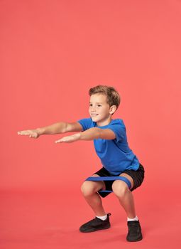 Cute male child looking away and smiling while doing squats with resistance exercise band against red background