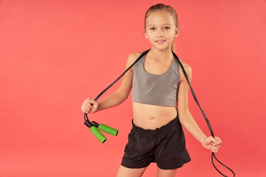 Adorable female child in sportswear looking at camera and smiling while holding jump rope