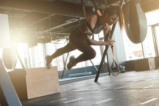 Pushing The Limits. Young athletic strong woman in sportswear doing TRX workout at industrial gym, exercising with fitness straps. Suspension training concept. Sport, wellness and healthy lifestyle