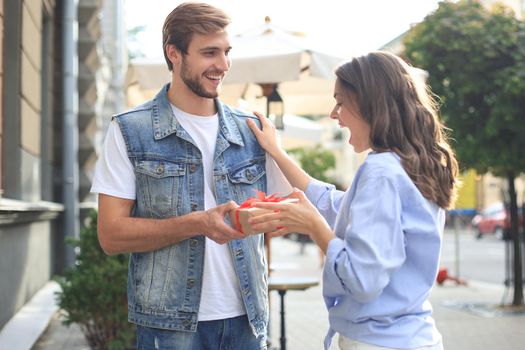 Astonished excited couple in summer clothes smiling and holding present box together while standing on city street