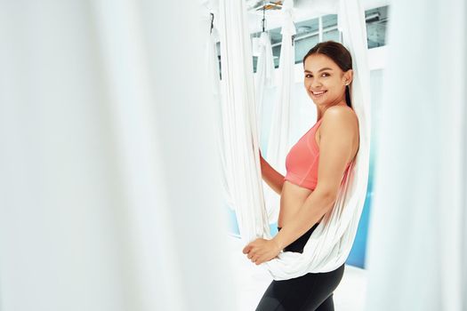 Young happy and beautiful woman in sportswear looking at camera and smiling while posing in bright fly yoga studio, holding white hammock suspended from the ceiling. Wellness and healthy lifestyle