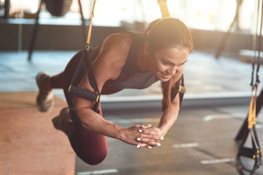 Working hard. Young athletic fitness woman in sportswear doing TRX workout at industrial gym, working out with fitness straps. Suspension training concept. Sport, wellness and healthy lifestyle