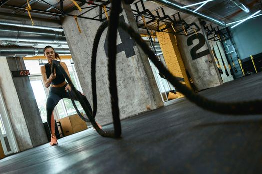 Crossfit workout. Young strong athletic woman wearing sport clothes exercising with battle ropes at gym, full length. Sport, training and healthy lifestyle