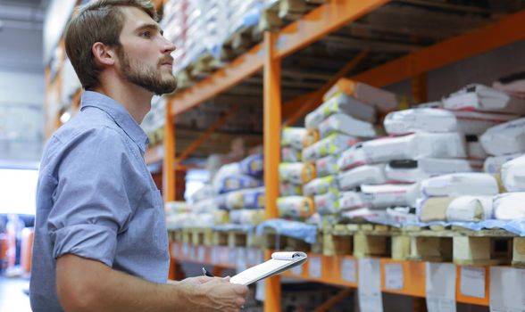 Young man shopping or working in a hardware warehouse standing checking supplies on his tablet
