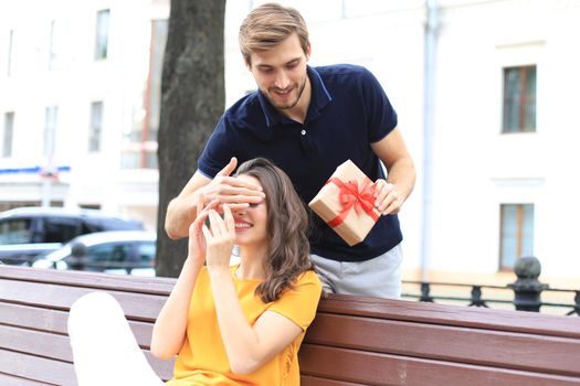 Image of charming excited couple in summer clothes smiling and holding present box together while sitting on bench