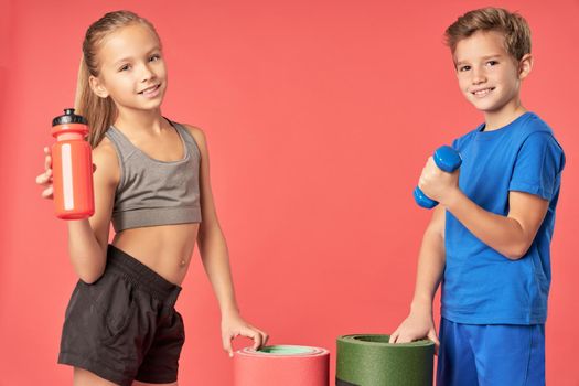 Adorable girl with bottle of water looking at camera and smiling while boy holding dumbbell and yoga mat