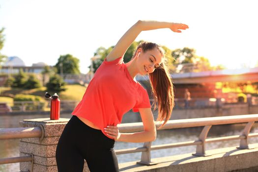 Image of beautiful young woman in sportive clothes stretching her body while doing workout near city riverfront in the evening