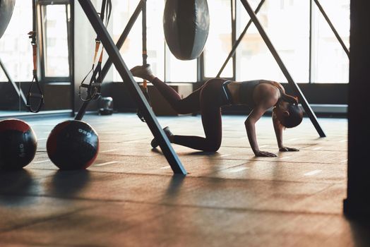 Side view of a young athletic woman in sports wear training legs with fitness trx straps, exercising at industrial gym. Sport, workout, wellness and healthy lifestyle