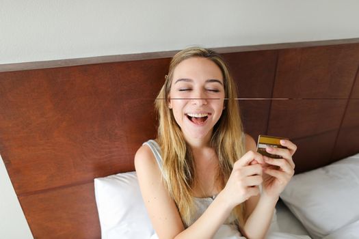 Young female person sitting in bed with gold debit card and smling, sitting in bed. Concept of payment thing and purchasing.