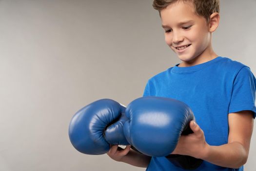 Adorable male kid looking at boxing gloves and smiling while standing against gray background