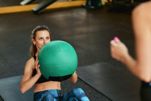 Young sportive woman in sportswear doing abs exercises exercising with fitness ball at gym with assistance of personal trainer, selective focus. Sport, training, wellness and healthy lifestyle