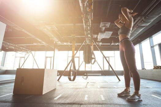 Prepering for workout. Side view of strong athletic woman in sportswear adjusting hair while standing at industrial gym, exercising with trx fitness straps. Sport and healthy lifestyle