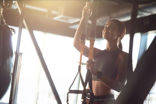 Young beautiful athletic woman in sports clothing adjusting trx fitness straps while standing at gym. Sport, workout, wellness and healthy lifestyle