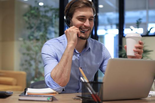 Cheerful young support phone male operator in headset, at workplace while using laptop, help service and client consulting call center concept.