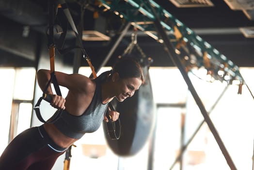 Side view of young beautiful fitness woman in sport wear doing TRX workout at industrial gym, doing push ups. Sport, training, wellness and healthy lifestyle