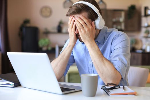 Unhappy frustrated young male holding head by hands sitting with laptop behind desk at home