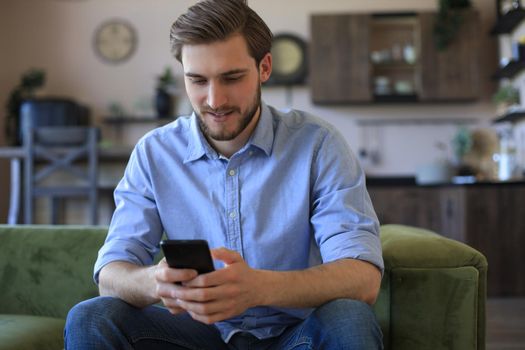 Attractive young man relaxing on a couch at home and using mobile phone for cheking social nets