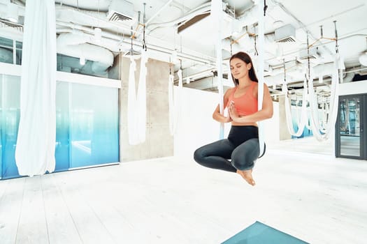 Ariel yoga. Young beautiful and calm woman in sportswear sitting in lotus pose in white hanging hammock and meditating, keeping eyes closed. Wellness and healthy lifestyle, harmony living